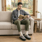 A man sits on a light beige sofa in a cozy room, reading a book. He is wearing a blue and gray plaid flannel shirt, olive green pants, white socks, and gray slip-on shoes.