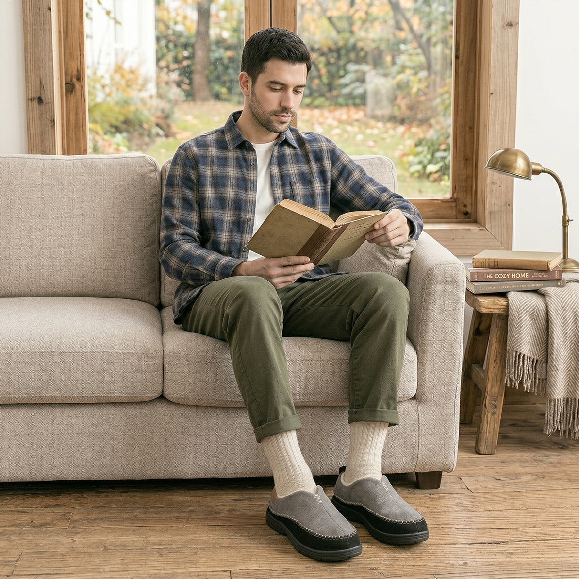 A man sits on a light beige sofa in a cozy room, reading a book. He is wearing a blue and gray plaid flannel shirt, olive green pants, white socks, and gray slip-on shoes.