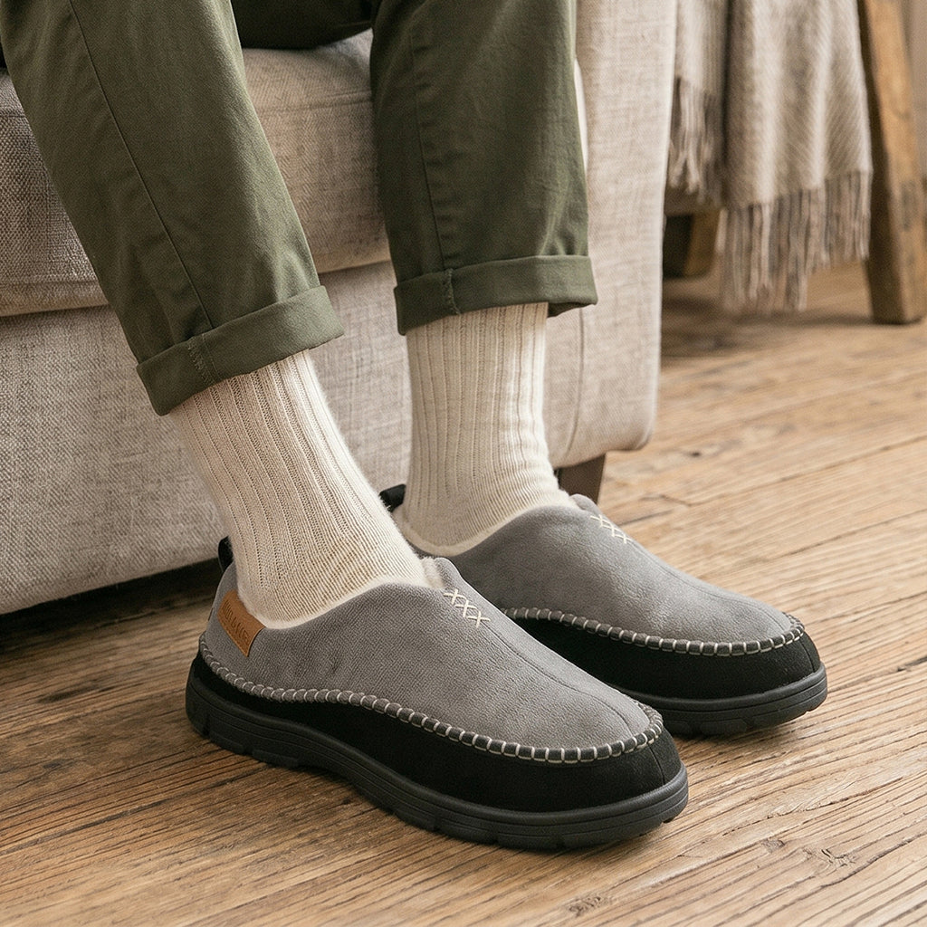 A person seated on a sofa wearing gray slip-on moccasin-style shoes with black soles, cream socks, and olive green pants, photographed in a home interior with a wood floor and beige couch.