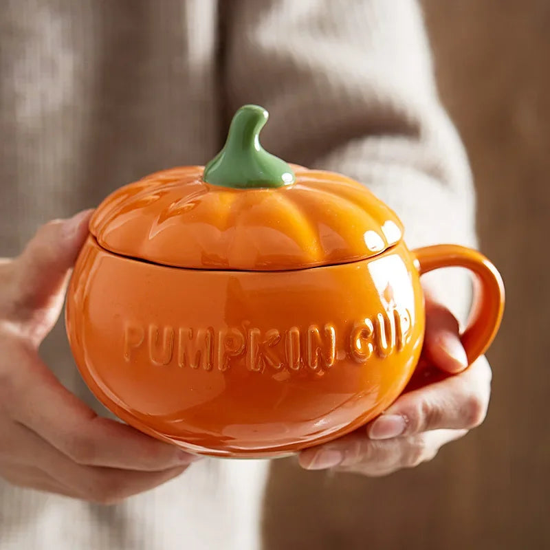 Orange pumpkin-shaped mug with a green lid held by hands against a neutral background