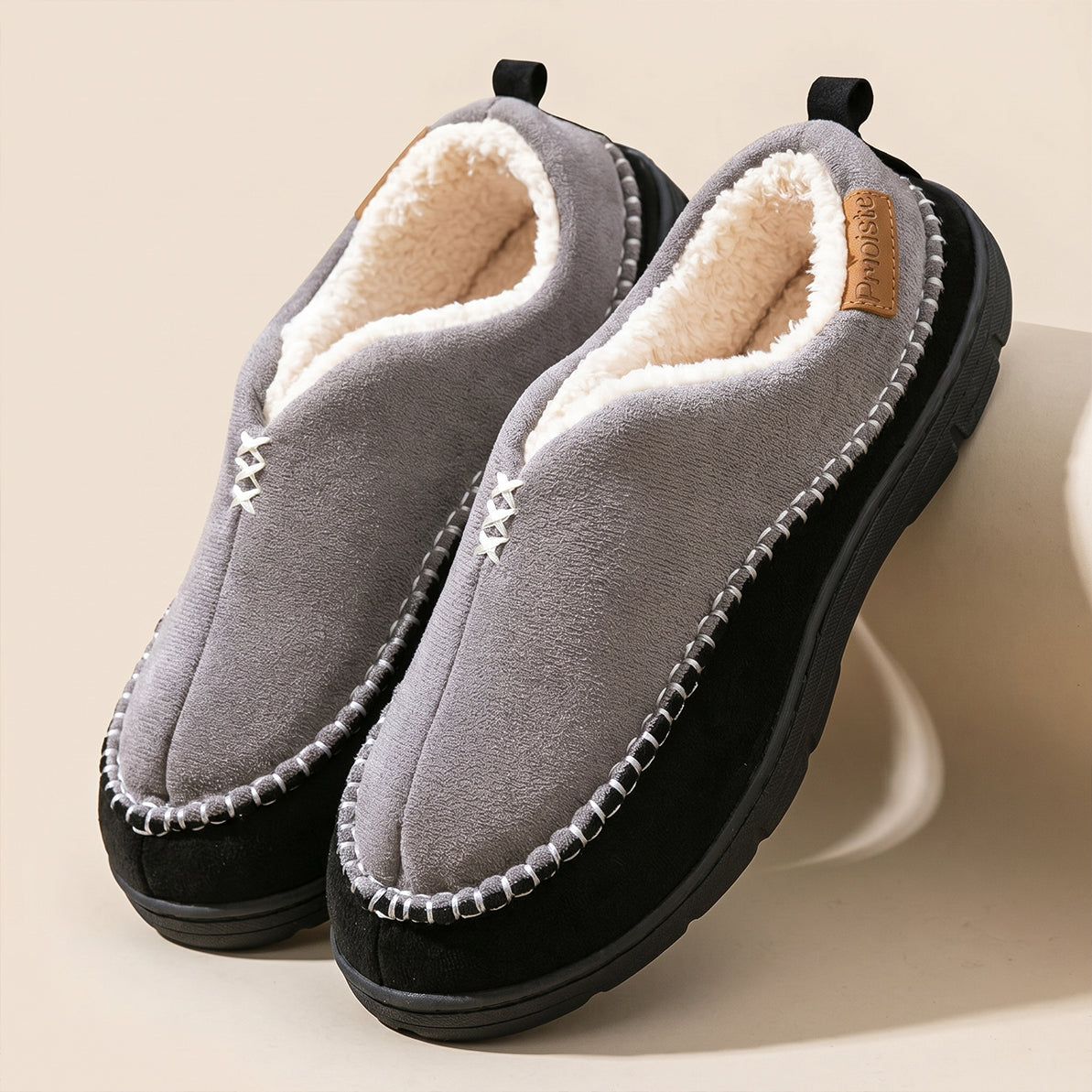 A pair of cozy slip-on winter slippers with a gray suede-like upper, cream sherpa lining, and black soles, displayed on a clean beige background.