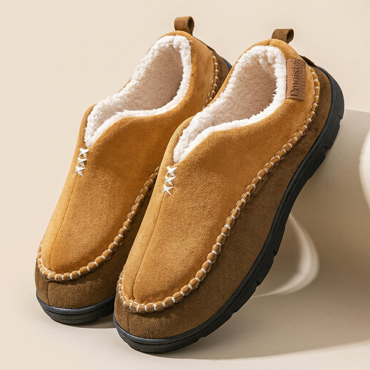 A pair of slip-on winter moccasin-style slippers in tan suede with a cream sherpa lining, decorative bead trim, and a black sole, displayed on a light beige background.