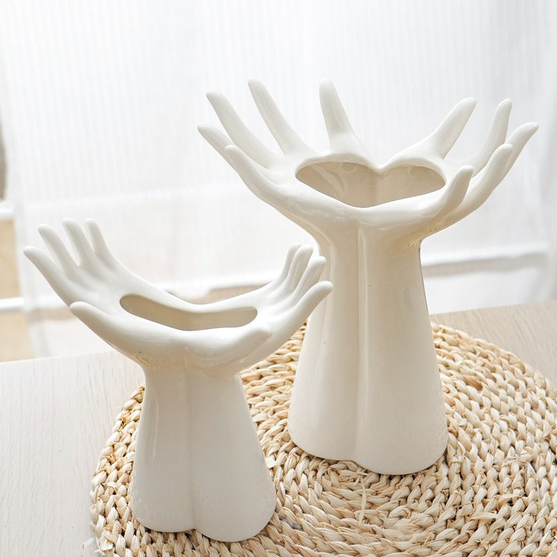 Two white ceramic hand-shaped candle holders with heart-shaped centers, displayed on a woven placemat on a light wooden table.