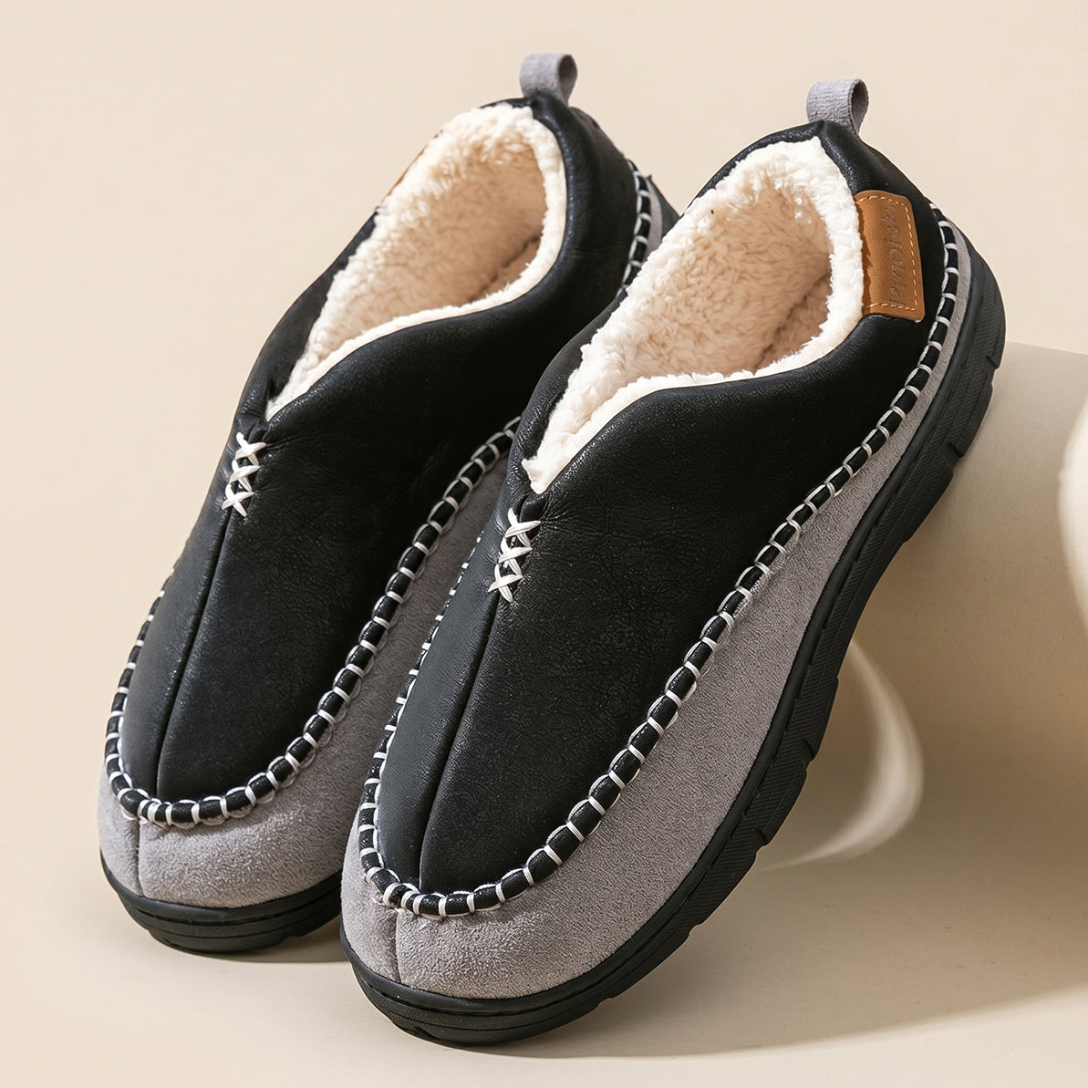 A pair of slip-on winter slippers with a black suede-like upper, light gray suede-like toe and heel sections, a cream sherpa lining, and decorative silver-tone stitching around the edges, displayed on a beige background.