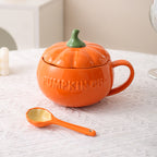 Orange pumpkin-shaped mug with a lid and matching spoon on a textured surface.