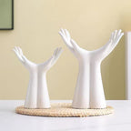 Two white ceramic hand-shaped decorative stands on a round woven mat, set on a white table against a beige wall.