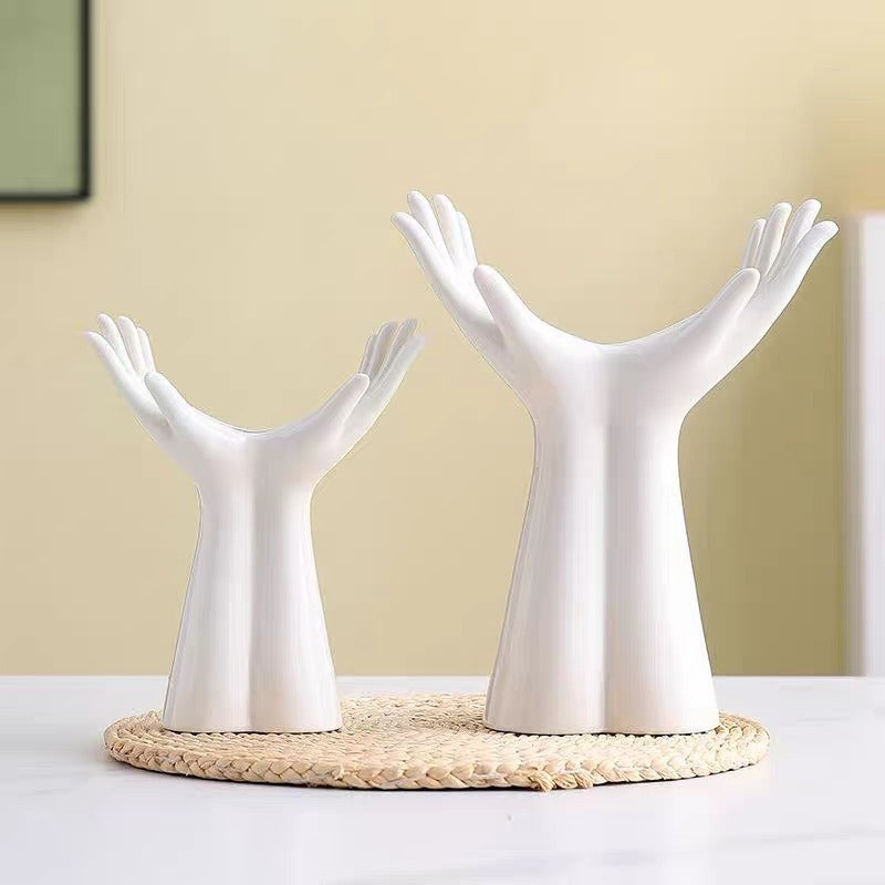 Two white ceramic hand-shaped decorative stands on a round woven mat, set on a white table against a beige wall.