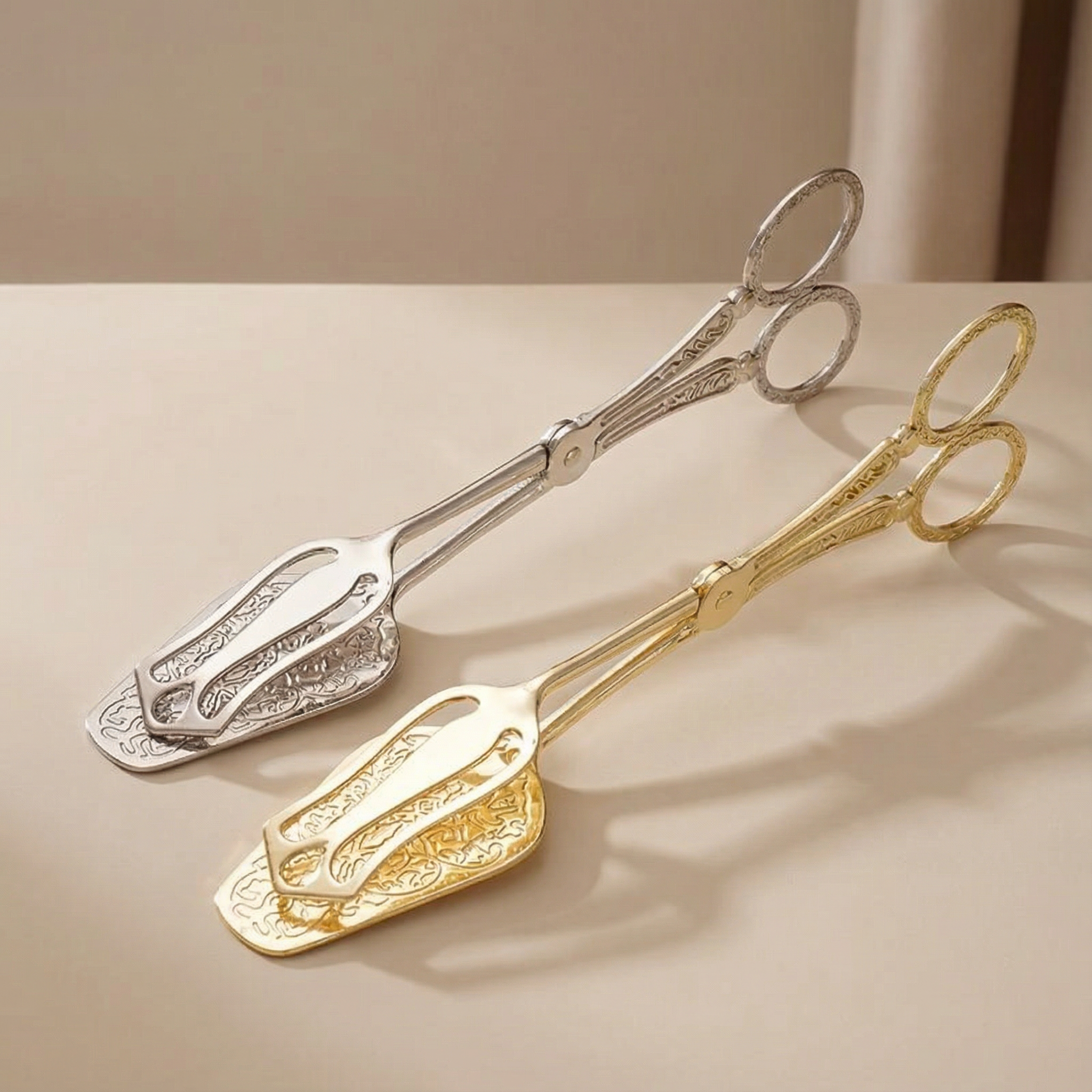 Two pairs of ornate tongs, one silver and one gold, on a beige surface.