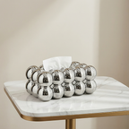 Decorative silver tissue holder with tissues on a marble table against a beige wall.