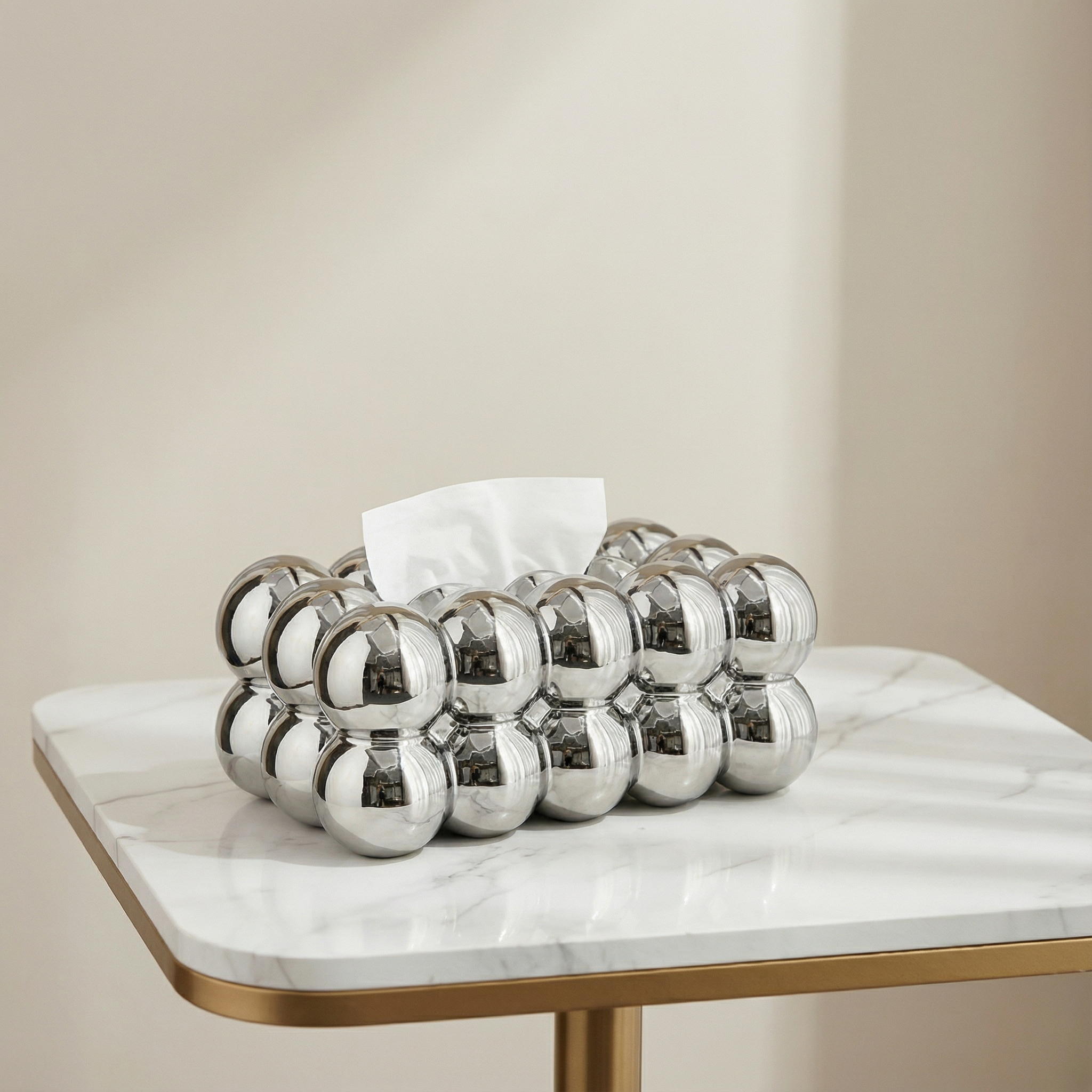 Decorative silver tissue holder with tissues on a marble table against a beige wall.