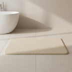 Beige bath mat on a tiled bathroom floor with a white sink in the background.