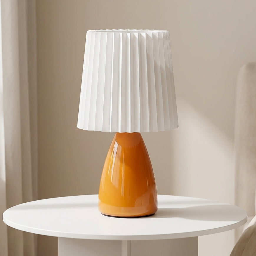 Table lamp with an orange base and white pleated shade on a round white table.