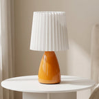 Table lamp with an orange base and white pleated shade on a round white table.
