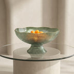 Decorative glass bowl with ribbed design on a pedestal, containing oranges, on a clear glass table against a neutral background.