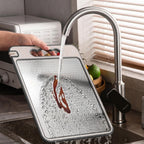 Person washing a cutting board under running water from a kitchen faucet.