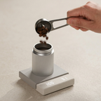 Hand pouring coffee beans into a container on a digital scale with a beige background