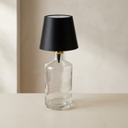 Clear glass bottle repurposed as a lamp with a black lampshade on a neutral background