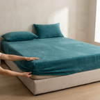 Person adjusting a teal fitted sheet on a bed in a minimalistic room.