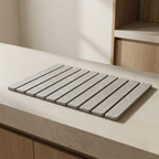 A rectangular gray slatted mat or tray with rounded corners, placed on a light beige countertop in a modern kitchen setting with wood cabinetry visible in the background.