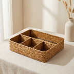 A rectangular woven tray with five compartments, made from natural tan seagrass or wicker, displayed on a light beige tablecloth next to a white ceramic vase with dried grasses.