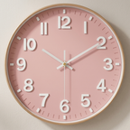 A round wall clock with a light pink face, large white Arabic numerals, white hands, and a thin light wood frame, shown against a plain light beige wall.