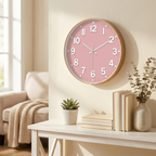 A round wall clock with a pink face and gold-toned frame hangs on a cream wall above a white console table styled with books, a small plant, and dried stems.