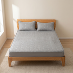 A gray mattress fitted with a striped gray and white mattress protector and two matching pillowcases, shown on a wooden bed frame in a bright, minimalist bedroom.
