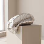 A sculptural, curved object covered in small reflective silver tiles, displayed on a beige pedestal in a softly lit interior setting.