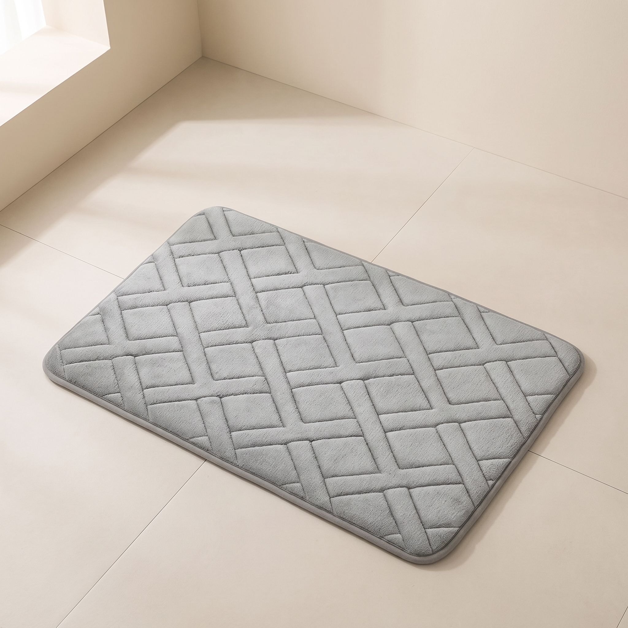 A light gray rectangular bath mat with a raised geometric basketweave pattern, shown on a beige tiled floor in a brightly lit room.