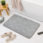 A gray rectangular bath mat with a raised basketweave pattern, styled in a bathroom setting with a white bathtub, beige slippers, a towel, and a small wooden stool.