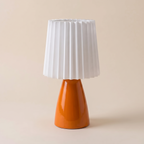 A modern table lamp with a glossy orange ceramic base and a white pleated fabric shade, set against a solid beige background.