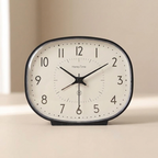 A HoneyTime analog desk clock with a rounded rectangular shape, featuring a cream-colored face, black numerals, and black hands, set against a minimalist beige background.