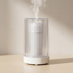 A white and clear ribbed cylindrical humidifier or diffuser sitting on a light wood surface against a warm beige wall, with visible mist rising from the top.