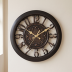 A large round wall clock with a dark brown frame and ornate cutout numerals, featuring gold-tone hands and a decorative central medallion, mounted on a light beige wall.