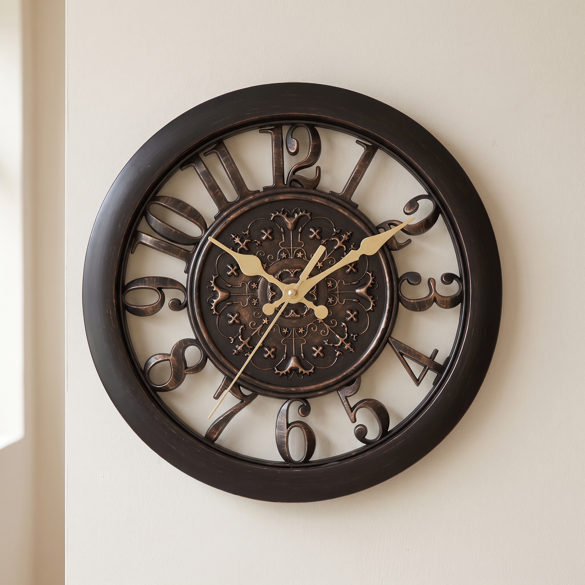 A large round wall clock with a dark brown frame and ornate cutout numerals, featuring gold-tone hands and a decorative central medallion, mounted on a light beige wall.