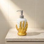 A bathroom soap dispenser with a white cylindrical bottle and a metallic gold hand-shaped base, set on a light marble countertop against a beige tiled wall.