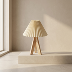 A pleated beige table lamp with a natural wood tripod base, displayed on a matching beige pedestal in a minimalist room setting with warm sunlight.