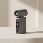 A modern dark gray personal fan with a digital display showing 100% and a silver button, standing on a light beige surface with soft shadows.