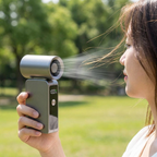 A woman outdoors holds a compact handheld fan with a cylindrical top and visible air jet, shown in profile against a blurred green park background.