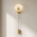 A modern wall-mounted light fixture with a circular backplate, warm white illuminated ring, slim vertical rod, and cylindrical gold-toned base, shown against a plain off-white wall with soft sunlight and shadows.