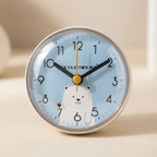 A round desk clock with a light blue face, black numerals, black hands, and a white polar bear illustration near the bottom. The clock has a silver metal rim and is photographed on a warm beige surface with a softly blurred background.