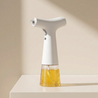 A modern electric oil spray bottle with a white T-shaped body and a clear ribbed base filled with golden oil, standing on a white surface against a beige background.
