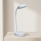 A modern, minimalist table lamp in a very light pastel blue-gray color, featuring a circular base and a flexible gooseneck arm ending in a flat, disc-shaped shade. It is shown resting on a light beige wooden surface against a matching beige wall, casting a soft shadow.