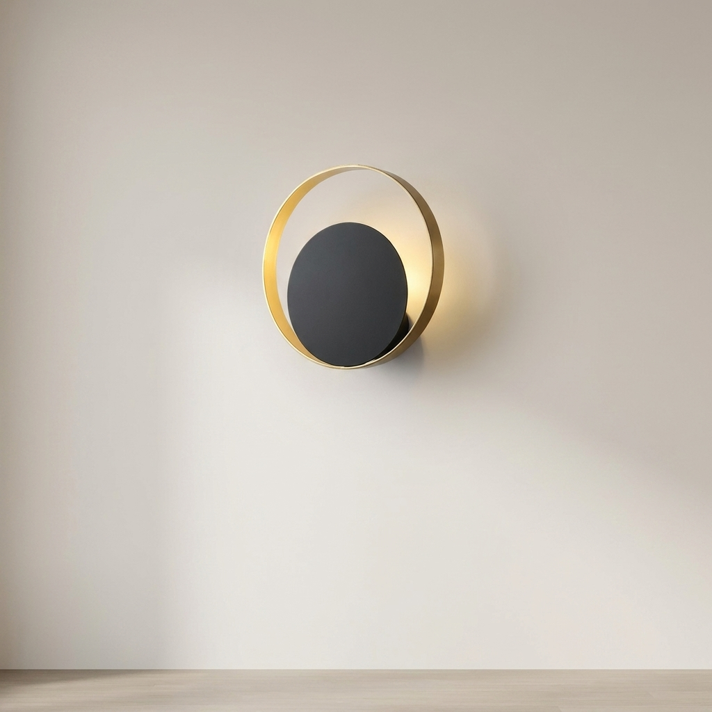 A modern wall sconce light fixture with a matte black circular center and a gold-toned circular frame, mounted on a light beige wall above a light wood floor.
