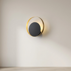 A modern wall sconce light fixture with a matte black circular center and a gold-toned circular frame, mounted on a light beige wall above a light wood floor.