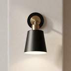 A modern wall sconce light fixture with a matte black cone-shaped shade, a black round wall plate, and a brushed gold neck, mounted on a textured off-white wall.