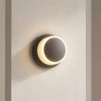 A modern circular wall sconce light fixture with a matte dark gray outer shade and a glowing warm white illuminated inner ring, mounted on a textured off-white wall.