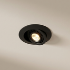 A round black recessed spotlight ceiling light fixture emitting a warm yellow glow, shown against a plain beige wall.