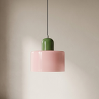 A modern pendant light with a glossy pink cylindrical shade and a green cylindrical accent above it, suspended by a thin black cord in a minimalist room with beige walls and a gray floor.