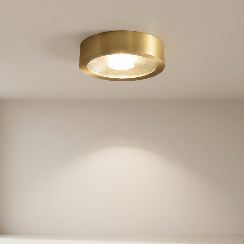 A modern flush-mount ceiling light fixture in brushed gold with a frosted glass diffuser, shown illuminated on a clean white ceiling and wall.